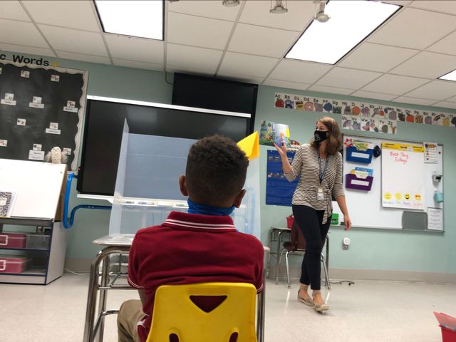 a boy sits at his desk while wearing mask and watching teacher through barrier